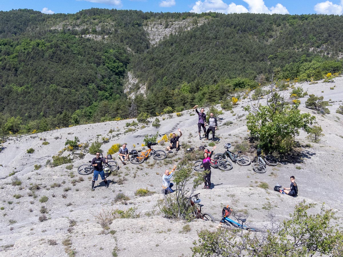 Group of mountain bikers resting on the grey marl slopes of the Terres Noires in Digne-les-Bains, surrounded by bikes and forested scenery
