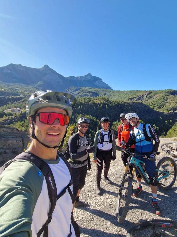Groupe de vttistes souriants posant pour une photo sur un sentier de montagne, avec un paysage de sommets et de forts en arrire-plan.