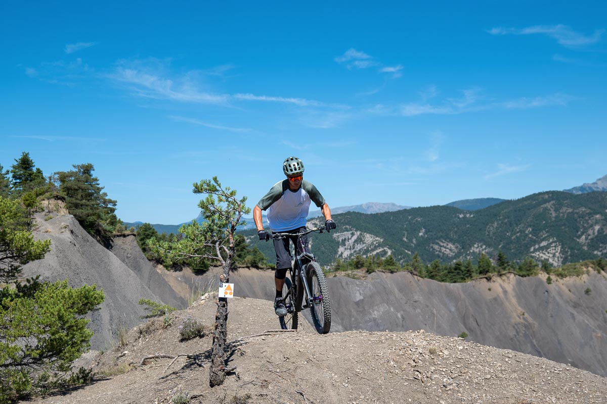 Vttiste descendant un sentier escarp en montagne sous un ciel bleu, portant un casque et des lunettes de protection.
